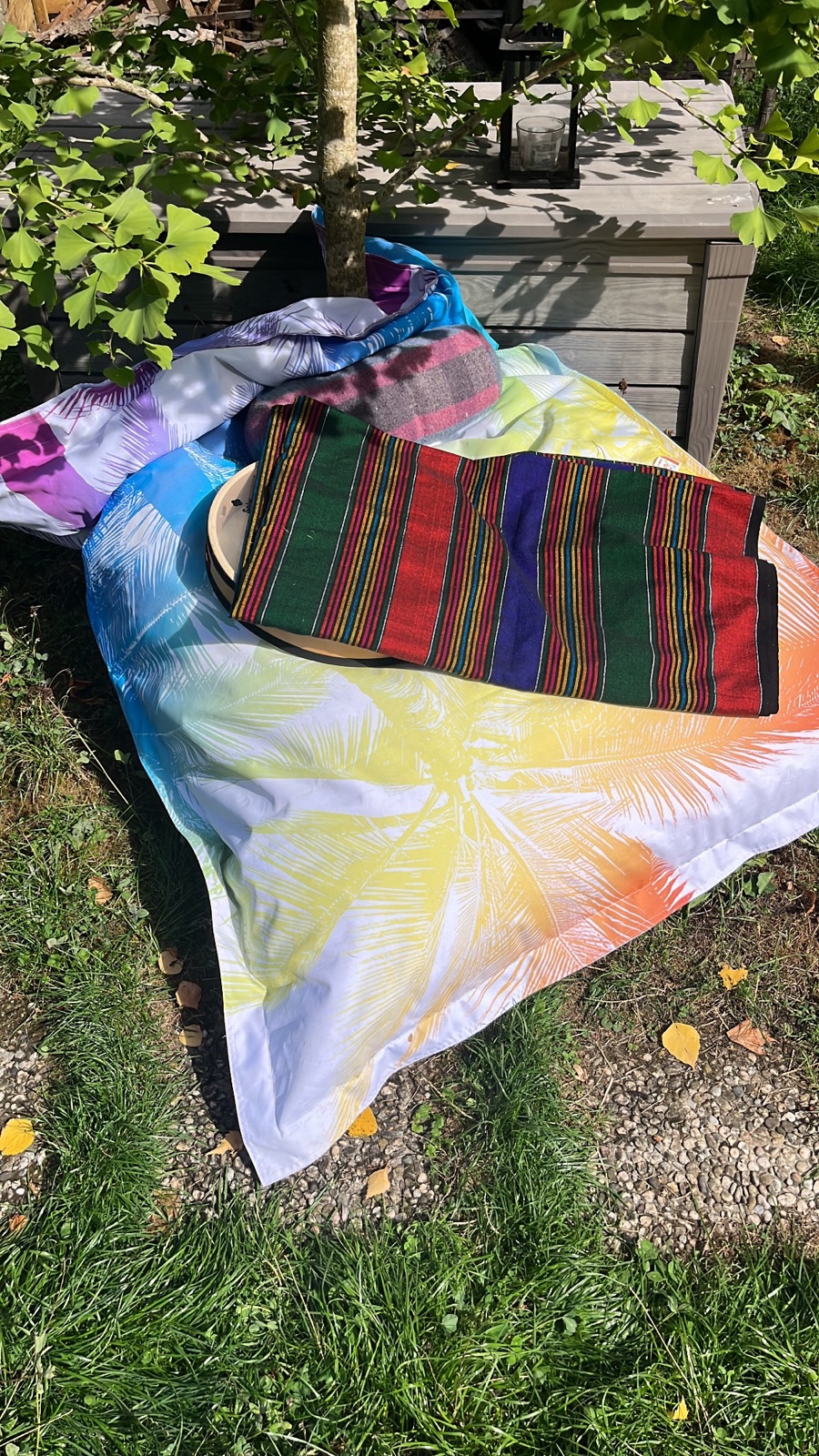 Bright cloths, a striped blanket and a drum piled around the base of a young tree in a sunlit garden.