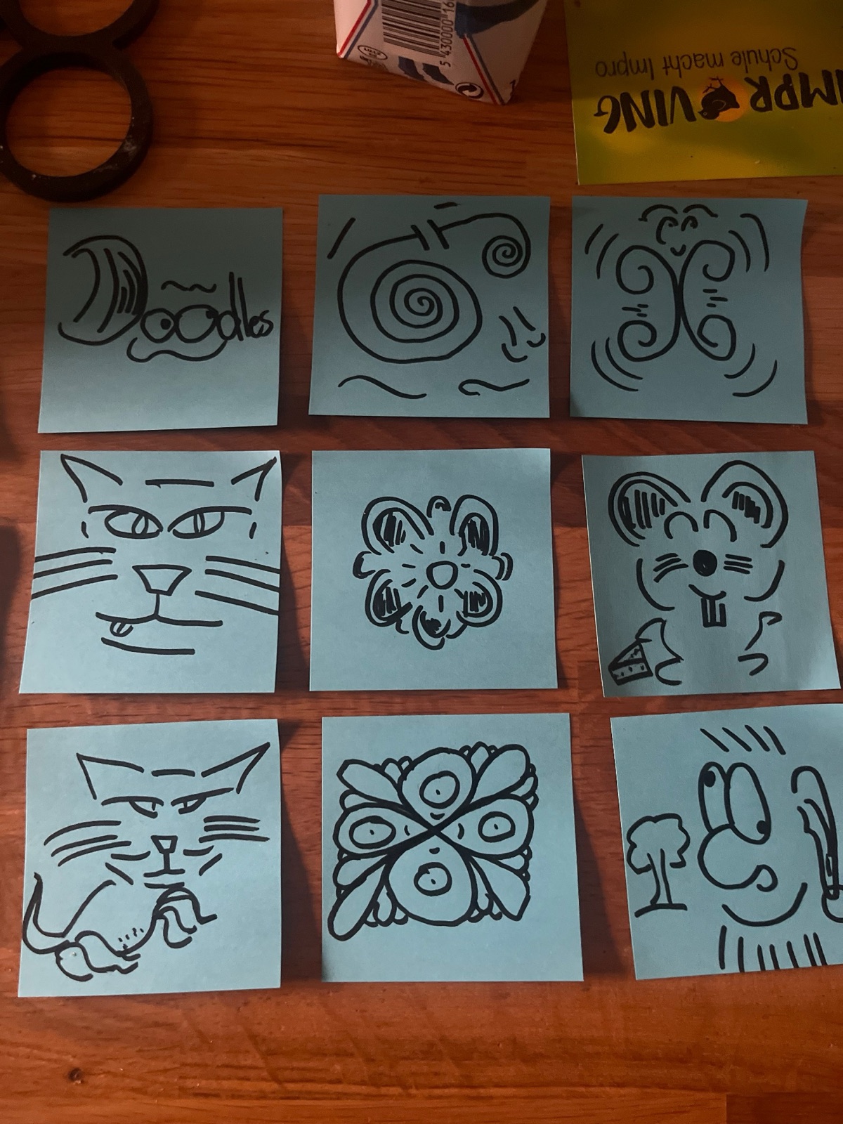 Nine small blue sticky notes laid out in a three-by-three grid on a wooden table, each covered in a bold black marker doodle: the word Doodles with eyes, spirals, a curling pattern, a cat's face, a flower, a mouse with cheese, a reclining cat, a butterfly, and a face beside a tree.