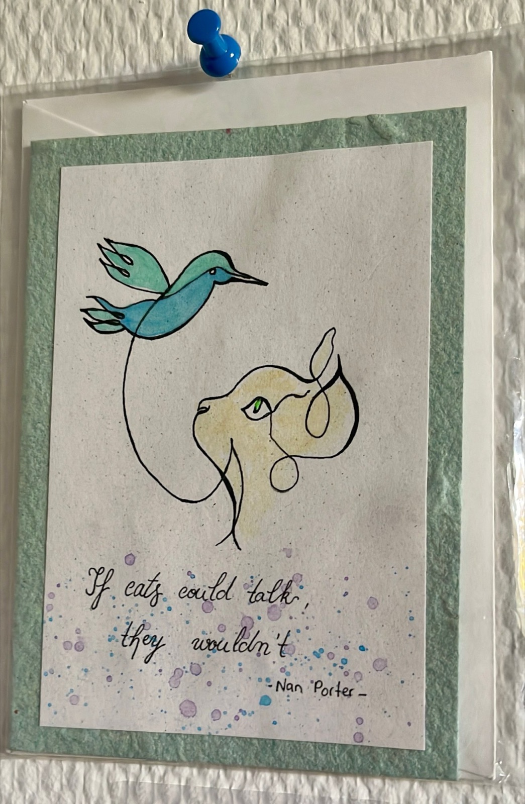 A pinned greeting card: an ink-and-watercolour line drawing of a turquoise bird in flight, its line running unbroken into the head of a pale ginger cat with one green eye. Beneath it, in handwriting: 'If cats could talk, they wouldn't.' Signed Nan Porter. The card is held to the wall with a single blue pin.